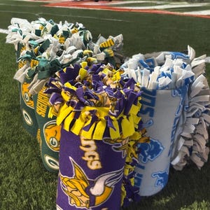 May include: Three cylindrical fleece blankets with knotted fringe edges, featuring team logos and colors. One is purple and yellow with a Viking graphic, one is green and yellow with a Packers logo, and one is light blue and white with a lion logo.