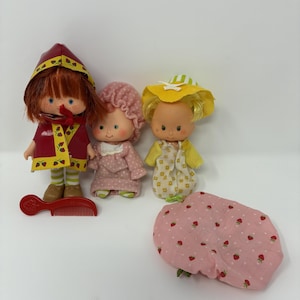 Peut inclure: Trois petites poupées avec des traits de visage peints et des cheveux enracinés. L'une porte une cape rouge à capuche avec des motifs de fraises, une autre un bonnet et une robe roses, et la troisième un chapeau jaune et une tenue à motifs. Un peigne en plastique rouge et un morceau de tissu rose à motifs de fraises.
