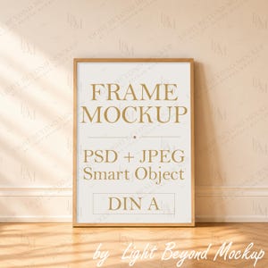 May include: A vertical rectangular wooden frame containing a white insert with gold text that reads FRAME MOCKUP, PSD + JPEG Smart Object, and DIN A. The frame has a light wood finish and is positioned against a light beige wall.