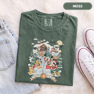 May include: A moss green t-shirt featuring a graphic print of Disney characters including Mickey Mouse, Minnie Mouse, Pluto, and Donald Duck in a snowy winter scene with a cabin, pine trees, and the text "Merriest Place Walt Disney World".