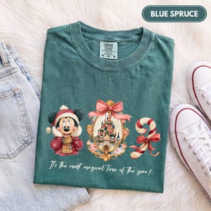 May include: A blue spruce t-shirt featuring a graphic of Minnie Mouse in a winter hat, a castle inside an ornate gold frame with a pink bow, and a candy cane. The text reads, It's the most magical Time of the year! The shirt has a crew neck.
