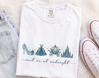 Könnte beinhalten: Ein weißes Kurzarm-T-Shirt mit einem Grafikdruck eines blauen Glasschuhs, eines blauen Ballkleides, einer Kürbiskutsche und eines Schlosses, akzentuiert mit goldenen Sternen. Der Text "meet me at midnight" ist in blauer Schreibschrift gedruckt.