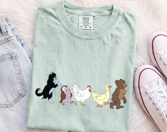 Könnte beinhalten: Ein hellgrünes T-Shirt mit einer Grafik, die eine schwarz-weiße Katze, eine kleine braune Eule, ein weißes Huhn, eine gelbe Ente und einen braunen Bären zeigt, die in einer Reihe laufen. Das T-Shirt hat ein Comfort Colors Etikett mit dem Text BAY.