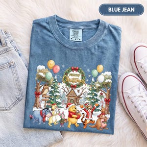 May include: A blue jean colored t-shirt featuring a graphic of Winnie the Pooh characters in a winter forest setting with a Christmas wreath and the text Merry Christmas. The shirt has a crew neckline and a Comfort Colors brand label at the neck.