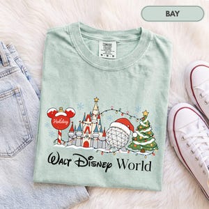 May include: A light green t-shirt featuring a graphic of a castle, a geodesic sphere wearing a Santa hat, a Christmas tree, and a Mickey Mouse-shaped ornament. The text reads Walt Disney World. The shirt has a Comfort Colors brand label.