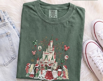 Könnte beinhalten: Ein moosgrünes T-Shirt mit einer Grafik eines Schlosses, umgeben von Feuerwerk und Luftballons in Mickey-Mouse-Form. Unter dem Schloss ist eine Gruppe von Figuren in Winterkleidung und Weihnachtsmützen von hinten zu sehen.