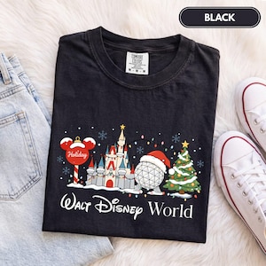 May include: A black t-shirt featuring a graphic of a holiday-themed castle, a geodesic sphere wearing a Santa hat, and a Christmas tree. The text reads Walt Disney World. The shirt has a Comfort Colors label at the neck and is shown with jeans and sneakers.