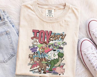 Könnte beinhalten: Ein elfenbeinfarbenes T-Shirt mit einem Grafikdruck der Toy Story-Figuren Woody, Buzz Lightyear, Jessie, Rex, Slinky Dog, Bullseye und Hamm. Der Text lautet Toy Story und enthält die Namen der Figuren sowie den Schriftzug Since 1995.