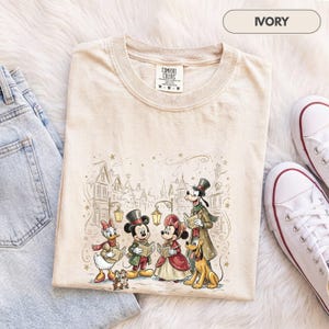 Könnte beinhalten: Ein elfenbeinfarbenes T-Shirt mit einem Grafikdruck von Disney-Figuren, darunter Mickey Maus, Minnie Maus, Donald Duck, Daisy Duck, Goofy und Pluto, die in einem Dorf Weihnachtslieder singen. Das T-Shirt hat einen Rundhalsausschnitt.