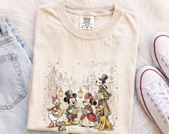 Op de afbeelding: Een ivoorkleurig t-shirt met een grafische print van Disney-figuren, waaronder Mickey Mouse, Minnie Mouse, Donald Duck, Daisy Duck, Goofy en Pluto, die kerstliedjes zingen in een dorp. Het t-shirt heeft een ronde hals.