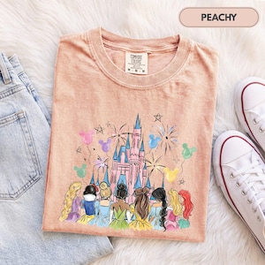Könnte beinhalten: Ein pfirsichfarbenes T-Shirt mit einer Grafik eines rosa Schlosses, umgeben von Feuerwerk und bunten Mickey-förmigen Luftballons. Unter dem Schloss befinden sich Illustrationen mehrerer Figuren mit langem Haar in verschiedenen Farben.