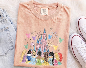 Könnte beinhalten: Ein pfirsichfarbenes T-Shirt mit einer Grafik eines rosa Schlosses, umgeben von Feuerwerk und bunten Mickey-förmigen Luftballons. Unter dem Schloss befinden sich Illustrationen mehrerer Figuren mit langem Haar in verschiedenen Farben.