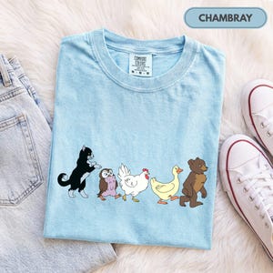 K&ouml;nnte beinhalten: Ein hellblaues T-Shirt mit einem Grafikdruck, der eine schwarz-wei&szlig;e Katze, eine kleine braune Eule, ein wei&szlig;es Huhn, eine gelbe Ente und einen braunen B&auml;ren zeigt, die in einer Reihe gehen. Das T-Shirt hat ein Comfort Colors Etikett und Chambray.
