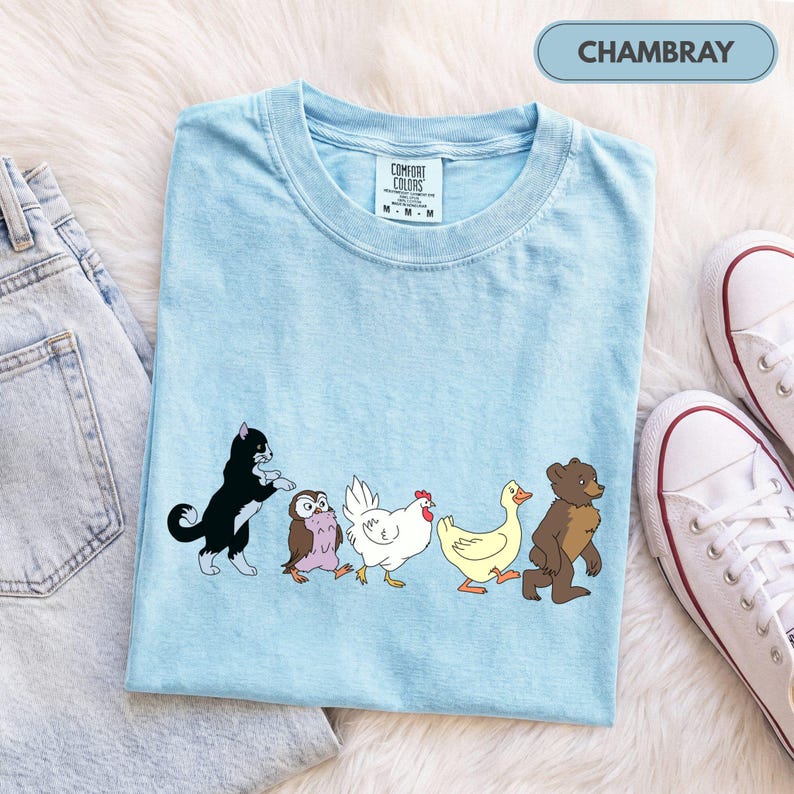 K&ouml;nnte beinhalten: Ein hellblaues T-Shirt mit einem Grafikdruck, der eine schwarz-wei&szlig;e Katze, eine kleine braune Eule, ein wei&szlig;es Huhn, eine gelbe Ente und einen braunen B&auml;ren zeigt, die in einer Reihe gehen. Das T-Shirt hat ein Comfort Colors Etikett und Chambray.