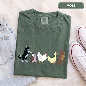 K&ouml;nnte beinhalten: Ein moosgr&uuml;nes T-Shirt mit einem Grafikdruck, der eine schwarz-wei&szlig;e Katze, eine braune Eule, ein wei&szlig;es Huhn, eine gelbe Ente und einen braunen B&auml;ren zeigt, die in einer Reihe laufen. Das T-Shirt hat einen Rundhalsausschnitt und ein Comfort Colors Etikett.