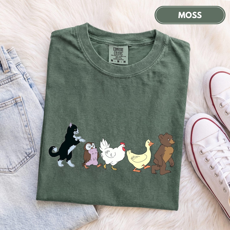 K&ouml;nnte beinhalten: Ein moosgr&uuml;nes T-Shirt mit einem Grafikdruck, der eine schwarz-wei&szlig;e Katze, eine braune Eule, ein wei&szlig;es Huhn, eine gelbe Ente und einen braunen B&auml;ren zeigt, die in einer Reihe laufen. Das T-Shirt hat einen Rundhalsausschnitt und ein Comfort Colors Etikett.