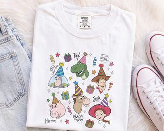 Könnte beinhalten: Ein weißes T-Shirt mit einem bunten Grafikdruck verschiedener Zeichentrickfiguren mit Partyhüten, darunter ein Dinosaurier, ein Cowboy, ein Weltraum-Ranger, ein Schwein, ein Pferd und ein Cowgirl, umgeben von Geschenkboxen, Sternen und Namen.
