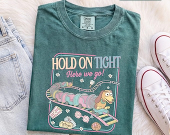 Könnte beinhalten: Ein blau-grünes Kurzarm-T-Shirt mit einer Grafik eines Dackels auf einer Achterbahn und dem Text "HOLD ON TIGHT Here we go!", umgeben von kleinen Blumen, einem Popcorn-Eimer, einer Sonnenbrille und einem Eintrittskarten-Abschnitt.