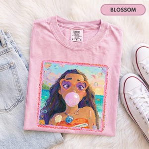 Könnte beinhalten: Ein hellrosa T-Shirt mit kurzen Ärmeln und einem quadratischen Grafikdruck einer Person mit langem dunklem Haar, die herzförmige Brillen trägt und eine rosa Kaugummiblase macht. Die Grafik zeigt eine Strandszene, eine Muschel und eine Halskette.