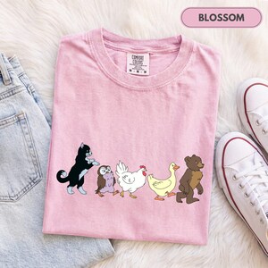 K&ouml;nnte beinhalten: Ein hellrosa T-Shirt mit einem Grafikdruck, der eine schwarz-wei&szlig;e Katze, eine kleine braune Eule, ein wei&szlig;es Huhn, eine gelbe Ente und einen braunen B&auml;ren zeigt, die in einer Reihe laufen. Das T-Shirt hat ein Comfort Colors Etikett am Innenkragen.