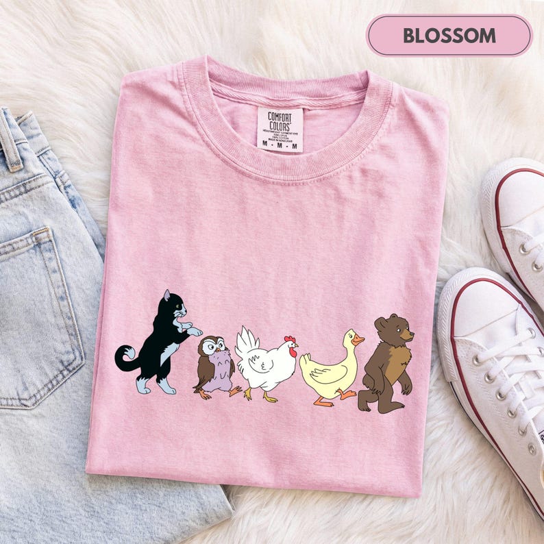 K&ouml;nnte beinhalten: Ein hellrosa T-Shirt mit einem Grafikdruck, der eine schwarz-wei&szlig;e Katze, eine kleine braune Eule, ein wei&szlig;es Huhn, eine gelbe Ente und einen braunen B&auml;ren zeigt, die in einer Reihe laufen. Das T-Shirt hat ein Comfort Colors Etikett am Innenkragen.