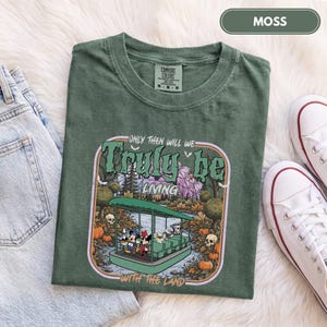 May include: A moss green short-sleeve t-shirt featuring a graphic print of a boat ride with cartoon characters. The text reads "ONLY THEN WILL WE TRULY BE LIVING WITH THE LAND". The shirt has a crew neckline and a tag indicating size M.