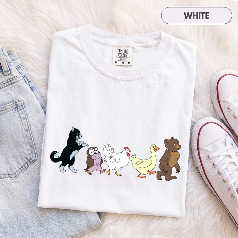 K&ouml;nnte beinhalten: Ein wei&szlig;es kurz&auml;rmeliges T-Shirt mit einem Grafikdruck von f&uuml;nf Tieren, die in einer Reihe laufen: eine schwarz-wei&szlig;e Katze, eine kleine braune Eule, ein wei&szlig;es Huhn, eine gelbe Ente und ein brauner B&auml;r. Das T-Shirt hat ein Comfort Colors Etikett.