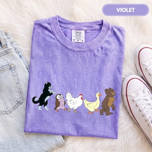 K&ouml;nnte beinhalten: Ein violettes kurz&auml;rmeliges T-Shirt mit einem Grafikdruck, der eine schwarz-wei&szlig;e Katze, eine braune Eule, ein wei&szlig;es Huhn, eine gelbe Ente und einen braunen B&auml;ren zeigt, die in einer Reihe laufen. Das T-Shirt hat einen Rundhalsausschnitt und ein Comfort Colors Etikett.