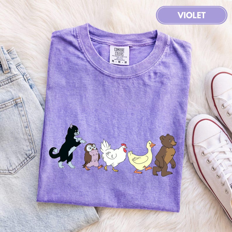 K&ouml;nnte beinhalten: Ein violettes kurz&auml;rmeliges T-Shirt mit einem Grafikdruck, der eine schwarz-wei&szlig;e Katze, eine braune Eule, ein wei&szlig;es Huhn, eine gelbe Ente und einen braunen B&auml;ren zeigt, die in einer Reihe laufen. Das T-Shirt hat einen Rundhalsausschnitt und ein Comfort Colors Etikett.