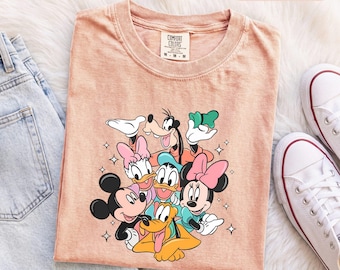Könnte beinhalten: Ein pfirsichfarbenes Kurzarm-T-Shirt mit einem Grafikdruck von Mickey Maus, Minnie Maus, Donald Duck, Daisy Duck, Goofy und Pluto. Das T-Shirt hat einen Rundhalsausschnitt und ein Markenetikett von Comfort Colors an der Innenseite des Kragens.