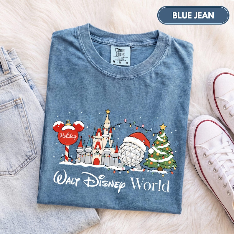 May include: A blue jean colored t-shirt featuring a graphic of a holiday-themed castle, a geodesic sphere wearing a Santa hat, and a Christmas tree, with the text Walt Disney World printed below in white script and sans-serif lettering.