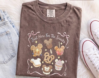 Könnte beinhalten: Ein braunes Kurzarm-T-Shirt mit einem Grafikdruck verschiedener Snacks in Form von Mickey-Mouse-Köpfen, darunter eine Waffel, ein Eis am Stiel, ein Liebesapfel, eine Brezel, ein Donut und ein Cupcake, mit dem Text Just Here For The Snacks.