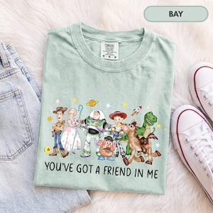 May include: A light green t-shirt featuring a graphic print of various animated characters including a cowboy, a space ranger, a potato figure, and a dinosaur. The text "YOU'VE GOT A FRIEND IN ME" is printed in black below the characters.