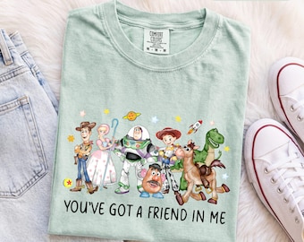 Könnte beinhalten: Ein hellgrünes T-Shirt mit einem Grafikdruck verschiedener animierter Figuren, darunter ein Cowboy, ein Weltraum-Ranger, eine Kartoffelfigur und ein Dinosaurier. Der Text "YOU'VE GOT A FRIEND IN ME" ist in Schwarz unter den Figuren aufgedruckt.