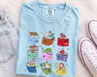 Könnte beinhalten: Ein hellblaues T-Shirt mit Rundhalsausschnitt und einem Grafikdruck von neun bunten, aufgeschlagenen Büchern, die jeweils eine andere Zeichentrickfigur und kleine Sternsymbole enthalten. Das Shirt hat ein Comfort Colors Etikett Größe M und die Farbe CHAMBRAY.