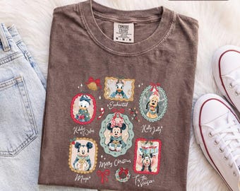 Könnte beinhalten: Ein espressofarbenes T-Shirt mit einem Grafikdruck von Disney-Figuren in verzierten Rahmen, darunter Mickey, Minnie, Donald und Pluto. Der Text umfasst 'Enchanted', 'Holiday Wishes', 'Holly Jolly', 'Merry Christmas' und 'Tis the season'.