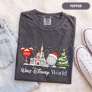 May include: A charcoal gray t-shirt featuring a graphic of a castle, a geodesic dome wearing a Santa hat, a Christmas tree, and a Mickey Mouse-shaped ornament. The text reads Walt Disney World. The shirt has a crew neck and short sleeves.