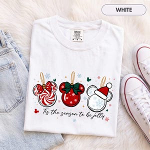 May include: A white short-sleeve t-shirt featuring a graphic of three Mickey Mouse-shaped Christmas ornaments. The ornaments include a peppermint swirl, a red bauble with a green bow, and a white bauble with a Santa hat. Text reads: Tis the season to be jolly.