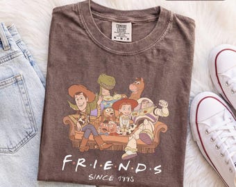 Könnte beinhalten: Ein braunes T-Shirt mit einer Grafik verschiedener animierter Figuren, die auf einem Sofa sitzen. Unter den Figuren steht der Text "F R I E N D S" und "SINCE 1995". Das T-Shirt hat einen Rundhalsausschnitt und ein Comfort Colors Markenetikett am Kragen.