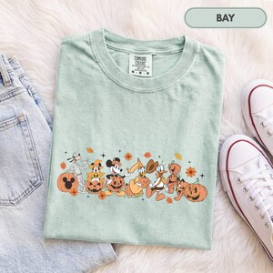 May include: A light green t-shirt featuring a graphic print of cartoon characters dressed in Halloween costumes with pumpkins. The shirt has a crew neckline and a white interior label with the text COMFORT COLORS, 100% COTTON, M-M-M.