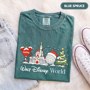 May include: A blue spruce colored t-shirt featuring a graphic of a castle, a geodesic sphere wearing a Santa hat, a Christmas tree, and a Mickey Mouse ornament. The text Walt Disney World is printed below the graphic in white script lettering.