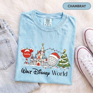 May include: A light blue t-shirt featuring a graphic of a castle, a geodesic sphere wearing a Santa hat, a Christmas tree, and a Mickey Mouse-shaped ornament. The text reads Walt Disney World. The shirt has a Comfort Colors brand label.