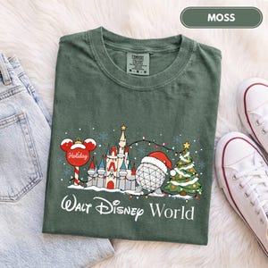 May include: A moss green t-shirt featuring a graphic of a castle, a geodesic sphere wearing a Santa hat, a Christmas tree, and a Mickey Mouse-shaped ornament. The text "Walt Disney World" is printed below the graphic in white lettering.