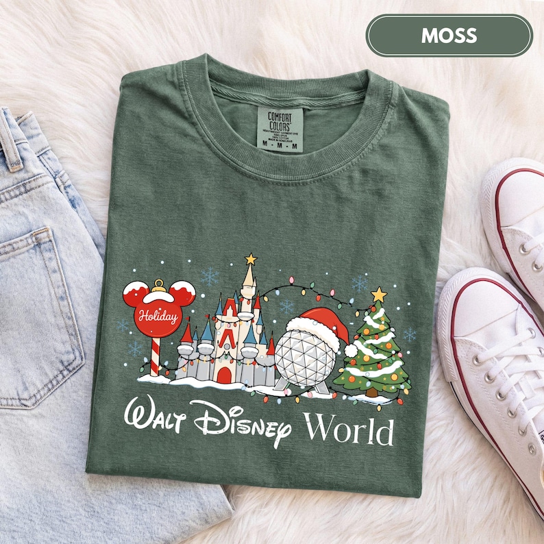 May include: A moss green t-shirt featuring a graphic of a castle, a geodesic sphere wearing a Santa hat, a Christmas tree, and a Mickey Mouse-shaped ornament. The text "Walt Disney World" is printed below the graphic in white lettering.