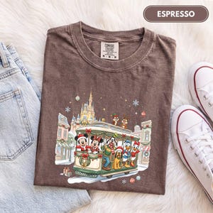 May include: An espresso brown t-shirt featuring a graphic print of Disney characters riding a holiday trolley in front of a castle. The characters include Mickey, Minnie, Goofy, Pluto, Donald, Daisy, and Chip and Dale. The text ESPRESSO is visible.