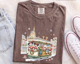 Könnte beinhalten: Ein espressobraunes T-Shirt mit einem Grafikdruck von Disney-Figuren, die in einer Weihnachtsbahn vor einem Schloss fahren. Zu sehen sind Mickey, Minnie, Goofy, Pluto, Donald, Daisy sowie Chip und Chap. Der Text ESPRESSO ist sichtbar.