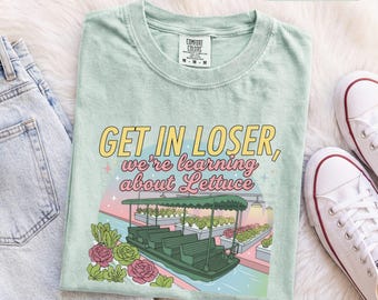 Könnte beinhalten: Ein hellgrünes T-Shirt mit einer Grafik eines Gewächshaus-Boots und dem Text "GET IN LOSER, we're learning about Lettuce" in Gelb und Pink. Das Design zeigt Illustrationen von Salatköpfen und rosa Blumen auf einem farbigen Hintergrund.