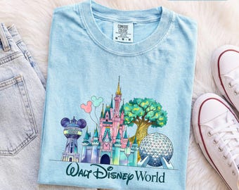 Könnte beinhalten: Ein hellblaues T-Shirt mit Rundhalsausschnitt und einem Grafikdruck, der ein Schloss, eine geodätische Kuppel, einen Wasserturm und einen Baum zeigt. Der Schriftzug Walt Disney World steht unter der Grafik. Das weiße Innenetikett zeigt Comfort Colors.