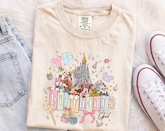 Könnte beinhalten: Ein elfenbeinfarbenes T-Shirt mit einem Grafikdruck von einem Schloss, Mickey Maus, Minnie Maus, Donald Duck, Daisy Duck, Goofy und Pluto. Der Text "birthday" ist in großen Blockbuchstaben gedruckt, "Girl" steht darunter in Schreibschrift.