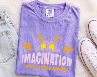 Könnte beinhalten: Ein violettes kurzärmeliges T-Shirt mit einer Grafik eines violetten Wesens mit gelben Augen und orangefarbenen Hörnern. Der Text lautet IMAGINATION in Hellrosa und ONE LITTLE SPARK in Orange. Das T-Shirt hat ein Comfort Colors Markenetikett.